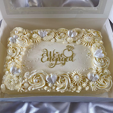 Engagement cake with white floral decorations, gold lettering, and decorative gemstones in a box. Perfect for celebrations.