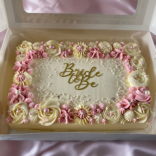 A beautifully decorated cake in a white box, featuring pink and white frosting roses and the phrase 