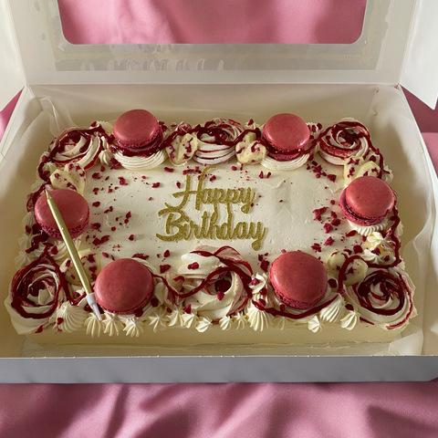 A beautifully decorated birthday cake with white frosting, pink macarons, and floral designs, featuring the words