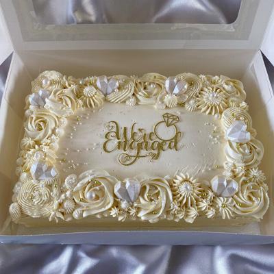 A beautifully decorated engagement cake with a creamy white frosting, adorned with intricate floral designs, sparkling hearts, and a golden inscription that reads